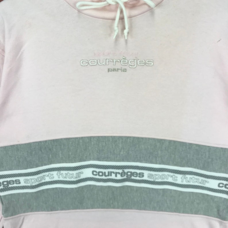 Op de afbeelding: Een lichtroze sweatshirt met een grijze horizontale streep over de borst. Het woord "courr&egrave;ges paris" is in lichtgrijs boven de streep geborduurd. De streep heeft de woorden "sport futur" en "courr&egrave;ges" in wit.