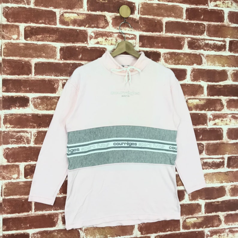 Op de afbeelding: Een lichtroze sweatshirt met lange mouwen en een grijze horizontale streep over de borst. De grijze streep heeft de merknaam "courr&egrave;ges" in het wit. De sweatshirt heeft een hoge halslijn met een trekkoord.