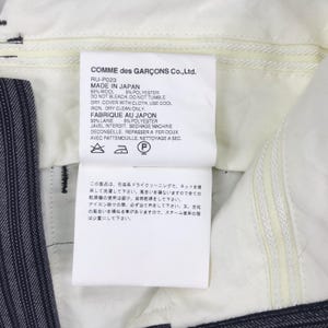 May include: White tag with black text detailing the care instructions for a pair of pants. The tag reads "COMME des GAR&Ccedil;ONS Co., Ltd. RU-P023 MADE IN JAPAN 95% WOOL 5% POLYESTER DO NOT BLEACH. DO NOT TUMBLE DRY. COVER WITH CLOTH, USE COOL IRON, DRY CLEAN ONLY. FABRIQUE AU JAPON 95% LAINE 5% POLYESTER JAVEL INTERDIT, SECHAGE MACHINE DECONSEILLE, REPASSER A FER DOUX AVEC PATTEMOUILLE. NETTOYAGE A SEC."