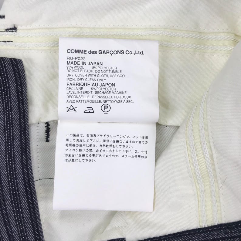 May include: White tag with black text detailing the care instructions for a pair of pants. The tag reads "COMME des GAR&Ccedil;ONS Co., Ltd. RU-P023 MADE IN JAPAN 95% WOOL 5% POLYESTER DO NOT BLEACH. DO NOT TUMBLE DRY. COVER WITH CLOTH, USE COOL IRON, DRY CLEAN ONLY. FABRIQUE AU JAPON 95% LAINE 5% POLYESTER JAVEL INTERDIT, SECHAGE MACHINE DECONSEILLE, REPASSER A FER DOUX AVEC PATTEMOUILLE. NETTOYAGE A SEC."