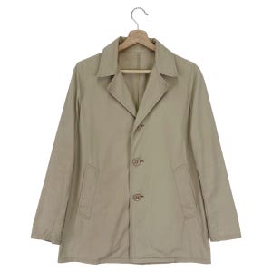 Puede incluir: Una chaqueta beige abotonada con cuello y dos bolsillos en la parte delantera. La chaqueta tiene tres botones en la parte delantera.