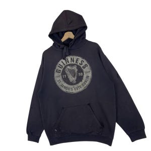 May include: Dark grey hooded sweatshirt with a kangaroo pocket and a drawstring hood. The front displays a circular graphic with the word "Guinness" and a harp design. The text "St. James's Gate Dublin" is also visible.