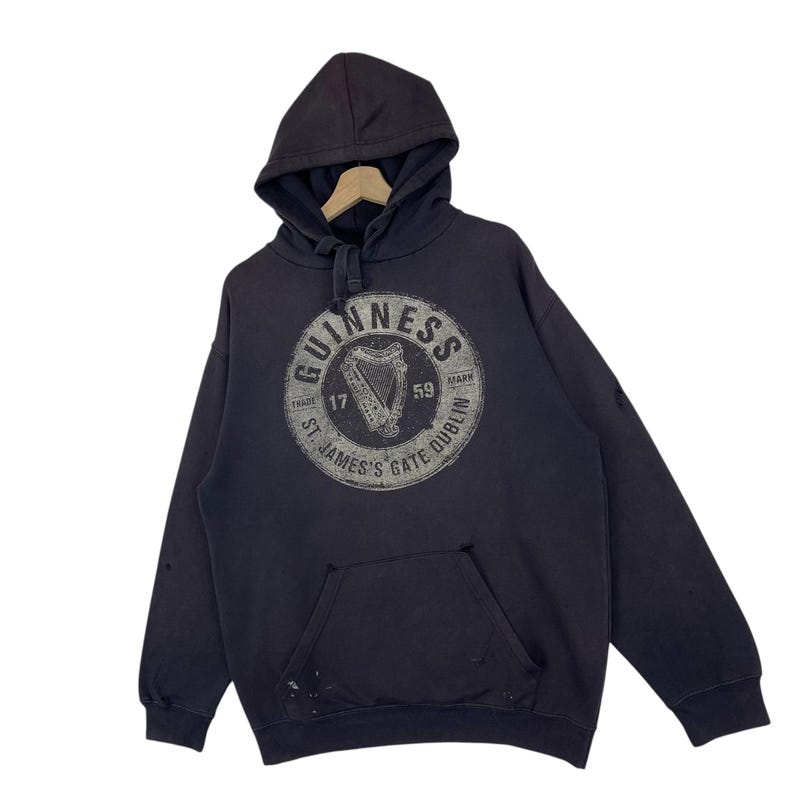 May include: Dark grey hooded sweatshirt with a kangaroo pocket and a drawstring hood. The front displays a circular graphic with the word "Guinness" and a harp design. The text "St. James's Gate Dublin" is also visible.