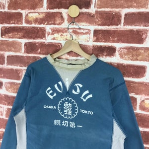 May include: A blue and beige long-sleeved sweatshirt with the word "Evisu" in a white circle with the words "Osaka" and "Tokyo" below it. The circle also contains Japanese characters and the words "新 功夫 第一".
