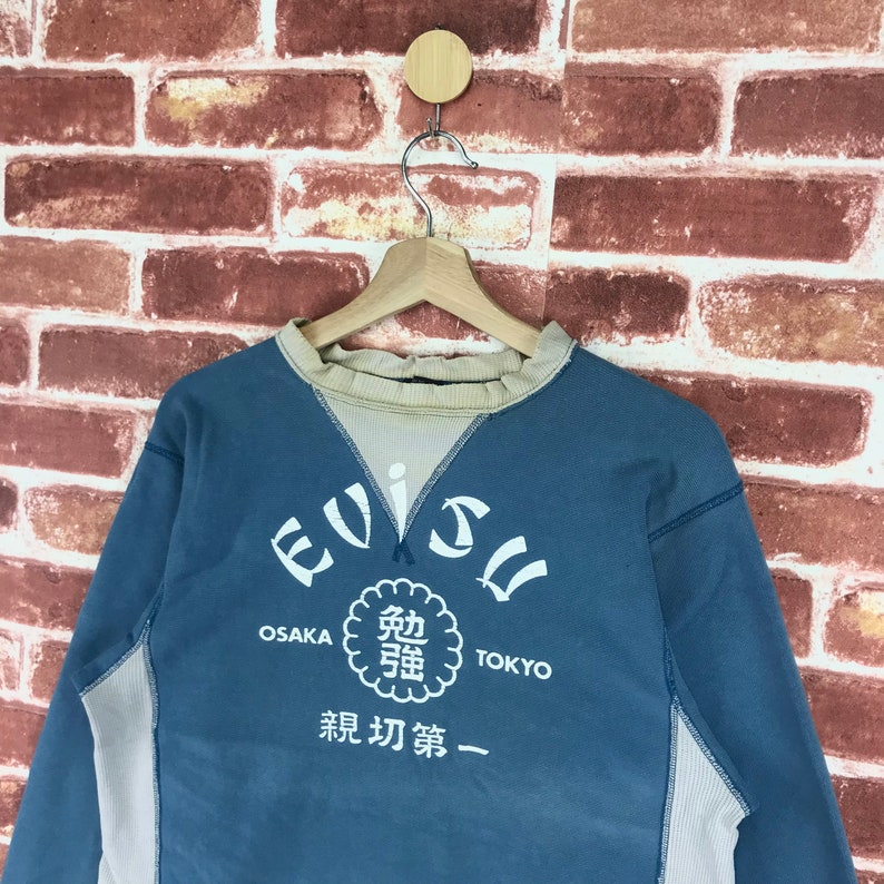May include: A blue and beige long-sleeved sweatshirt with the word "Evisu" in a white circle with the words "Osaka" and "Tokyo" below it. The circle also contains Japanese characters and the words "新 功夫 第一".