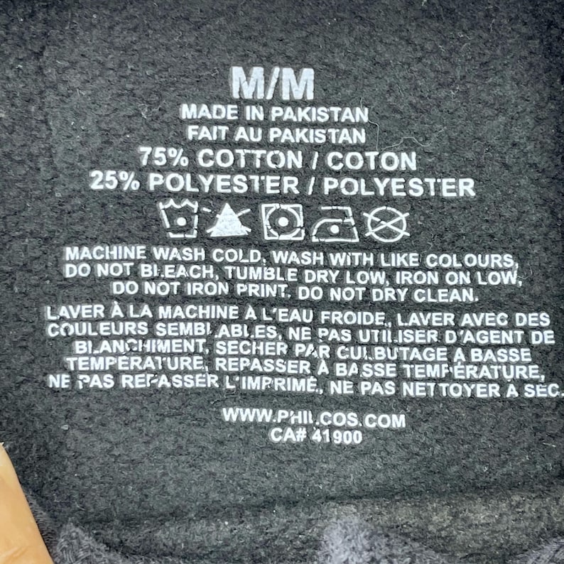 May include: A black garment label with white text detailing fabric composition and care instructions. The label includes the text "M/M", "Made in Pakistan", and washing symbols. The website address and CA number are also visible.