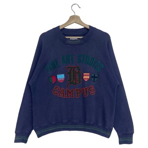 May include: A navy blue sweatshirt with the words "FINE ART STUDIOS CAMPUS" in green and red lettering. The sweatshirt has a crew neck and long sleeves. There are several crests with different designs on the front.