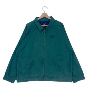 Puede incluir: Una chaqueta verde con cremallera con un logotipo bordado en azul en el pecho. La chaqueta tiene un cuello, puños y dobladillo de canalé.