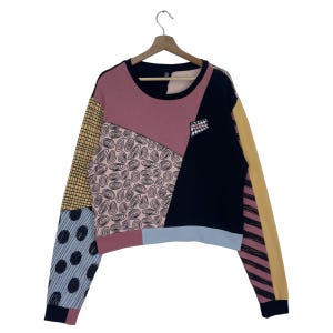 May include: A patchwork long-sleeved sweatshirt with a round neck. The design features various patterns and colours, including pink, black, yellow, and blue. The sleeves have different patterns, such as polka dots and stripes. The sweatshirt is hanging on a wooden hanger.