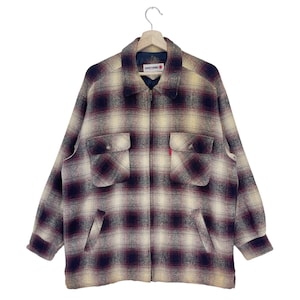 Op de afbeelding: Een bruin, geel en rood geruit flanellen jack met een ritssluiting. Het jack heeft twee grote zakken aan de voorkant en een klein rood label met de tekst "Lucky Strike" aan de linkerkant van de borst.