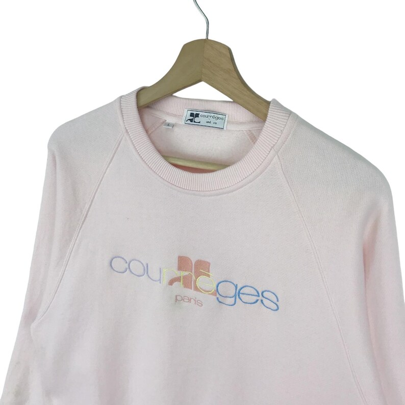 K&ouml;nnte beinhalten: Ein rosa Sweatshirt mit einem mehrfarbigen "Courr&egrave;ges Paris" Logo auf der Vorderseite.
