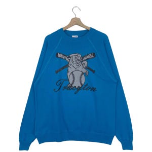 Puede incluir: Una sudadera azul con un gráfico de un pantera sosteniendo una pelota de béisbol y murciélagos cruzados. El texto "Truxton" está impreso debajo del gráfico.