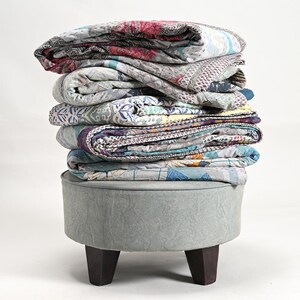 May include: A stack of colorful patchwork quilts folded on top of a light blue round ottoman with wooden legs.