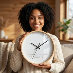 May include: A round wall clock with a light wood frame and a white face. The clock has black hands and the text "HE HAS MADE EVERYTHING BEAUTIFUL IN ITS TIME." ECCLESIASTES 3:11. The clock is being held by a person.