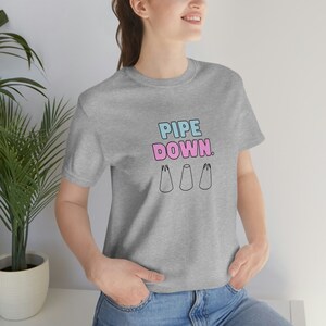 Peut inclure: Un t-shirt gris avec le texte "PIPE DOWN." en lettres roses et bleues. Trois poches à douille roses et bleues stylisées se trouvent sous le texte.