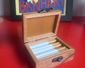Puede incluir: Una caja de madera con tapa abatible y cierre de metal. La caja está abierta y contiene una fila de cigarrillos blancos.