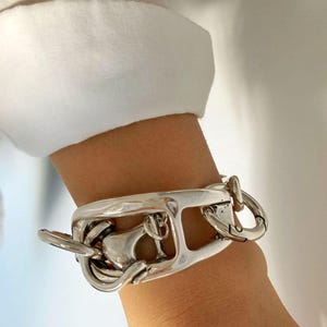 Peut inclure: Bracelet argenté au design unique, avec un élément rectangulaire central et une série de maillons et de boucles interconnectés. Le bracelet est porté au poignet, avec une manche de chemise blanche en arrière-plan.
