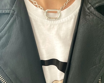 May include: A silver chain necklace with a large, oval-shaped clasp. The necklace is worn by a person wearing a white t-shirt and a black leather jacket.