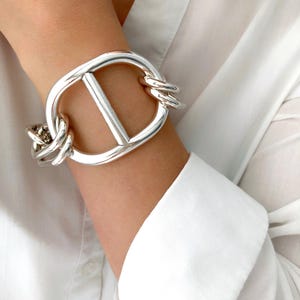 Peut inclure: Un bracelet argenté avec une pièce centrale rectangulaire et des maillons de chaîne. Le bracelet est porté sur un poignet, contrastant avec une chemise blanche à manches longues. Design audacieux, pour bijoux de mode.