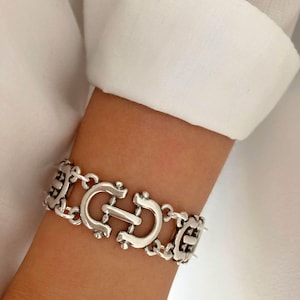 Puede incluir: Pulsera de plata con un diseño de cadena de eslabones y un gran cierre de 'C' entrelazado y adornado.