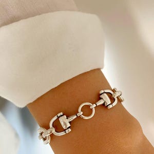 Op de afbeelding: Zilverkleurige armband met een paardenbit-ontwerp. De armband bestaat uit meerdere bitvormige schakels die door kleine ringen zijn verbonden. De sieraden worden om de pols gedragen, met een witte mouw op de achtergrond. Een klassiek accessoire geïnspireerd op paardensport.