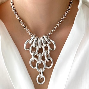 Può includere: Una collana argentata con una catena spessa e un gruppo di grandi maglie ovali ad incastro. La collana è indossata su una camicia bianca, evidenziando il suo design audace e la lucentezza metallica. Un accessorio di gioielleria.