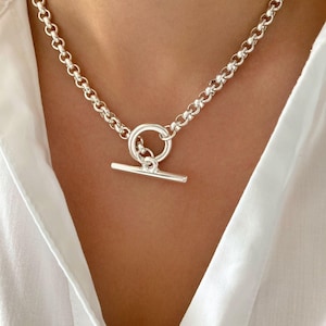 Peut inclure: Un collier argenté avec un fermoir à bascule. Le collier présente une chaîne à maillons arrondis et un fermoir circulaire avec une fermeture à barre. Le bijou est porté sur une chemise blanche.