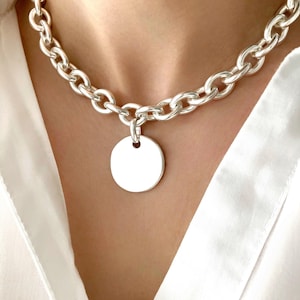 Peut inclure: Un collier en chaîne argentée avec un grand pendentif rond. La chaîne est épaisse et massive, et le pendentif est lisse et uni.