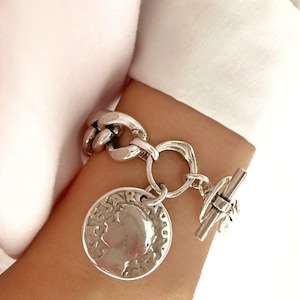 Könnte beinhalten: Silbernes Kettenarmband mit einem großen Münzenanhänger, der ein Profil einer Person zeigt. Die Münze trägt die Aufschrift "CESAR".
