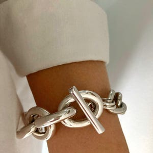 Peut inclure: Un bracelet en chaîne argentée épaisse avec un grand fermoir en forme de T.