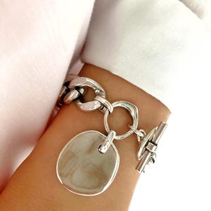 Puede incluir: Un brazalete de plata con una cadena grande y gruesa y un dije redondo y plano. El brazalete tiene un cierre de palanca.