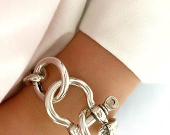 May include: A silver bracelet with a large, chunky, interlocking link design. The bracelet has a clasp that resembles a nautical shackle.