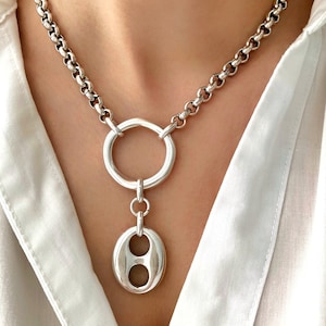 Peut inclure: Un collier en chaîne argent avec un grand pendentif rond orné d'un motif à double cercle.
