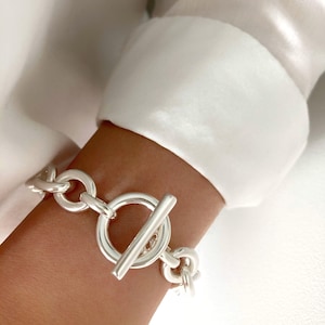 May include: A silver chunky chain bracelet with a large toggle clasp. The bracelet is made of thick, linked chains and has a polished finish.
