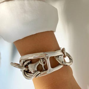 Peut inclure: Un bracelet en argent avec un design épais et ouvert. Le bracelet a un fermoir composé de plusieurs anneaux entrelacés.