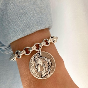 Könnte beinhalten: Ein silbernes Armband mit einem Münzanhänger. Der Anhänger zeigt das Profil einer Frau und die Inschrift "REPUBLIQUE FRANCAISE". Das Armband hat ein Kettendesign. Am Handgelenk ist ein blauer Hemdsärmel zu sehen.