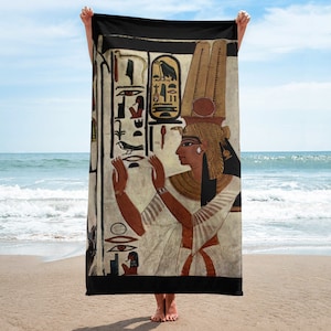 Puede incluir: Una toalla de playa negra con una imagen colorida de una reina egipcia que lleva un alto tocado. La reina está representada con los brazos levantados en un gesto de ofrenda. El fondo es una pared blanca con jeroglíficos.