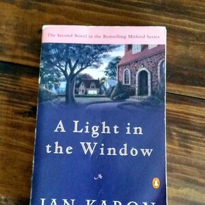 May include: A book cover with a blue background and the title "A Light in the Window" in white text. The author's name, "JAN KARON", is in white text below the title. The book is the second novel in the bestselling Mitford series and the author of "Out to Canaan".