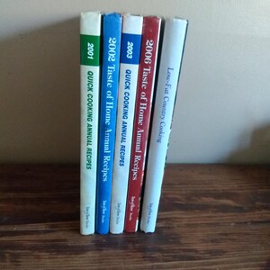 May include: A collection of cookbooks, including "2001 Quick Cooking Annual Recipes," "2002 Taste of Home Annual Recipes," "2003 Quick Cooking Annual Recipes," and "2006 Taste of Home Annual Recipes." The books are arranged vertically, with varying spine colors.