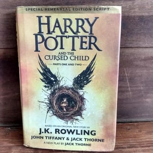 May include: A copy of "Harry Potter and the Cursed Child" book. The cover features the title in large, black letters, with a nest and bird wings graphic. The author's name, J.K. Rowling, is also visible. The book is a special rehearsal edition script.