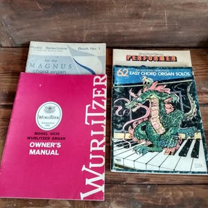 May include: A collection of vintage music books and manuals. Includes a red Wurlitzer Owner's Manual, a book of Magnus chord organ music, and a book titled "62 Easy Chord Organ Solos" with a dragon illustration. The items are on a wooden surface.