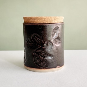 May include: A cylindrical ceramic canister with a dark brown, glossy glaze and a textured, embossed botanical leaf pattern on the exterior. The container features a flat, light brown cork lid that sits flush with the top rim of the vessel.