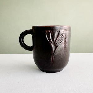 Peut inclure: Une tasse en céramique brun foncé avec un corps arrondi et une anse latérale unique. La surface extérieure présente un motif de feuille botanique subtilement en relief sur le devant, avec une finition mate et texturée sur toute la pièce.