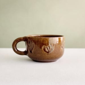 Può includere: Una tazza in ceramica marrone con finitura lucida e manico arrotondato. La superficie esterna presenta un motivo in rilievo ripetuto di fiori stilizzati a forma di ventaglio che circondano il corpo della tazza, creando un aspetto materico.
