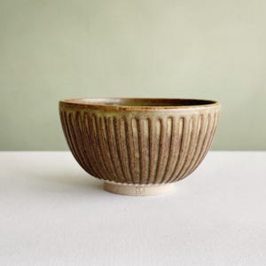 Peut inclure: Un bol en céramique de forme arrondie avec un extérieur texturé présentant des rainures verticales. Le bol possède une glaçure tachetée brune et beige, un intérieur lisse et une petite base circulaire non émaillée au centre du fond.
