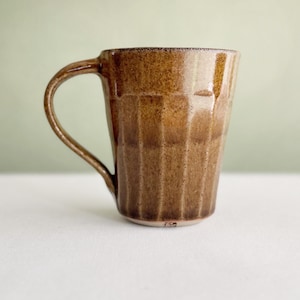 Peut inclure: Une tasse en céramique avec un vernis brun moucheté brillant et une texture nervurée verticale. La tasse présente une anse incurvée sur le côté gauche et une petite marque de signature sombre près de la base. La finition montre des nuances.