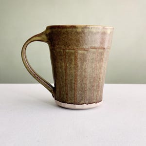 Peut inclure: Une tasse en céramique avec un émail moucheté vert olive et brun, présentant des lignes verticales indentées sur le corps, une anse incurvée et une base blanche lisse non émaillée. La tasse a un bord légèrement évasé et un aspect terreux.