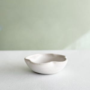 Peut inclure: Un petit bol en céramique blanche, façonné à la main, doté d'un bord irrégulier et ondulé. Le bol présente une finition mate lisse et un bassin peu profond et arrondi, offrant une forme simple et organique avec des indentations subtiles.