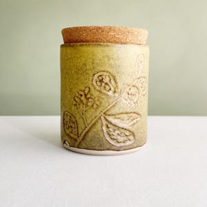 Może przedstawiać: Cylindryczny ceramiczny pojemnik z nakrapianą oliwkowozieloną glazurą oraz wypukłym, roślinnym wzorem liści i łodyg. Pojemnik jest zwieńczony okrągłą, jasnobrązową pokrywką z korka, która ściśle przylega do górnego otworu naczynia.