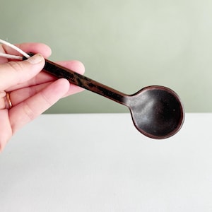 Peut inclure: Une petite cuillère en céramique brun foncé avec un bol rond et un long manche fin. La surface présente une finition mate texturée avec de subtiles variations de couleur. Une boucle en ficelle claire est fixée au bout du manche.